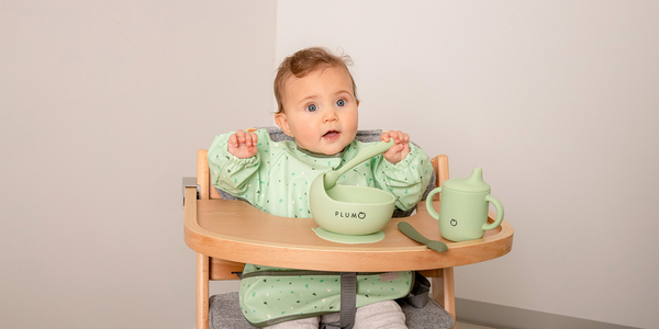 cute baby in a hi chair with plum brand feeding smock bowl and cup